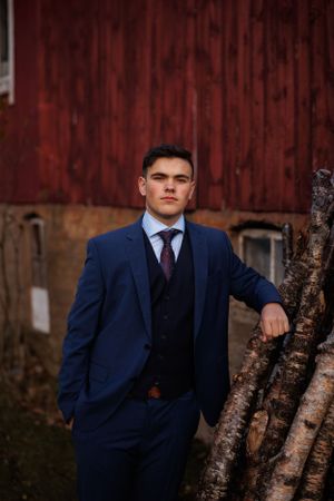 A person in a navy blue three-piece suit poses against a red barn wall with birch trees at sunset.