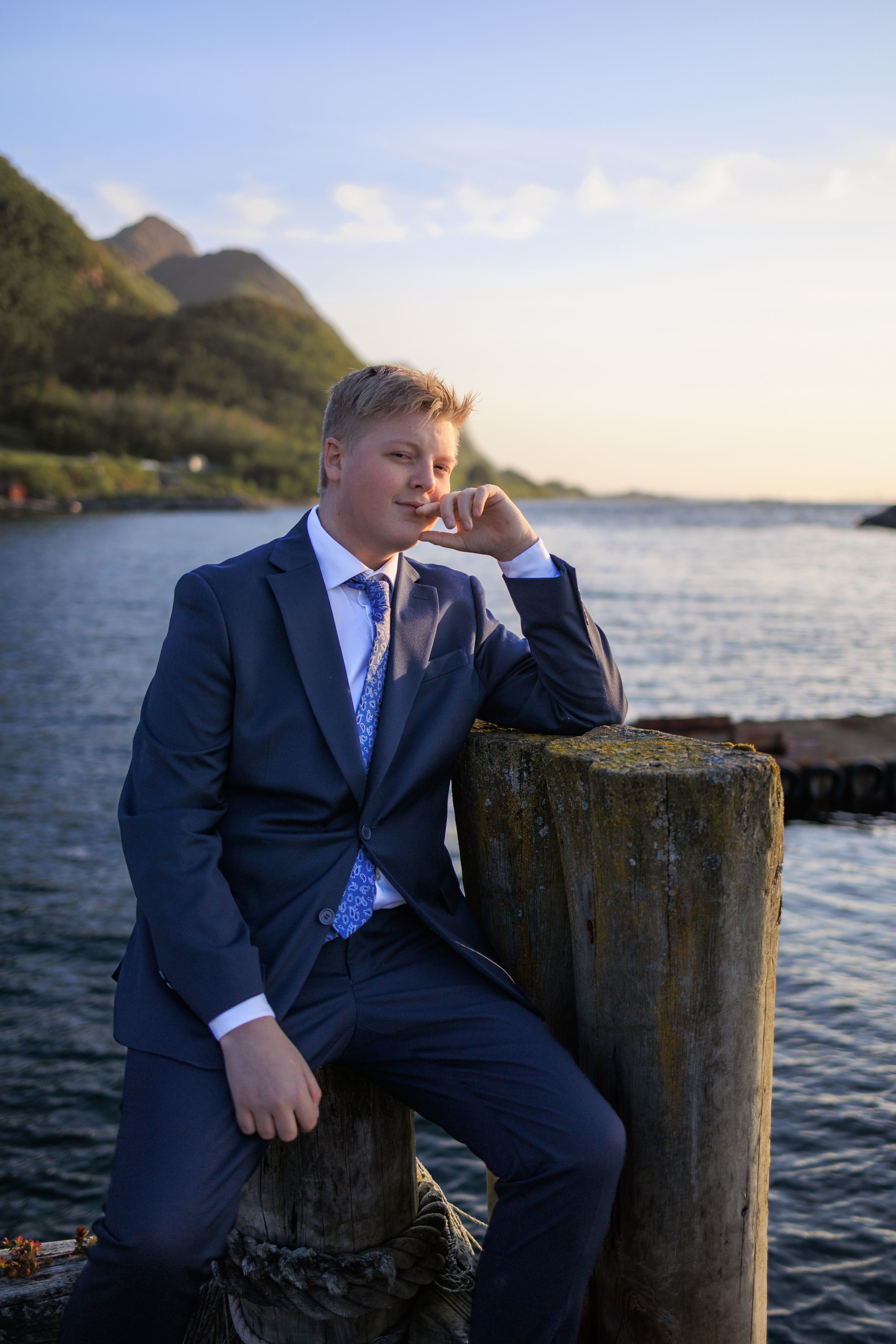 Well-dressed person in navy suit poses on coastal rocks at sunset with mountains and ocean in background.