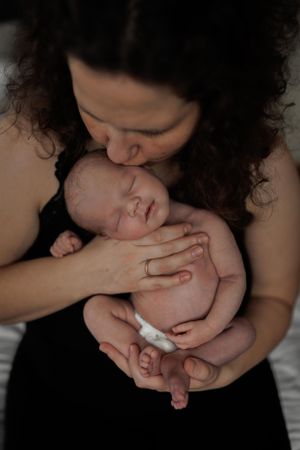A mother with curly dark hair tenderly kisses and holds her sleeping newborn baby against her chest.