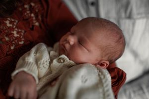 A sleeping newborn baby wearing white clothing rests peacefully against a red blanket in soft lighting.