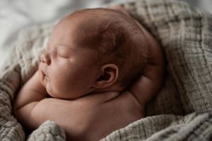 A sleeping newborn baby wrapped in a soft gray linen blanket lies peacefully on their side.