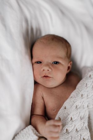 A small baby rests on soft white bedding while partially covered with a knit blanket.