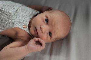 Sweet baby wearing a gray knitted sweater lays on bed while gently sucking their thumb.