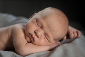A peaceful newborn baby sleeps serenely on soft grey blankets in a portrait-style photograph.