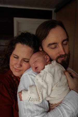 A newborn baby in a white outfit sleeps peacefully while being held by its parents.