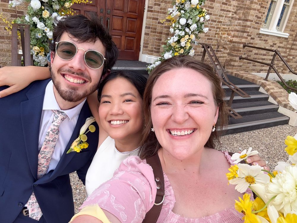 Three people smiling together outdoors in front of stone building with floral decorations and wooden door.