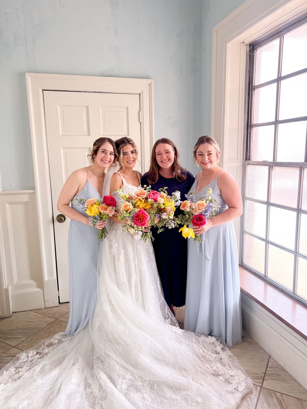 Bride in lace gown poses with bridesmaids holding colorful bouquets in light-filled room.