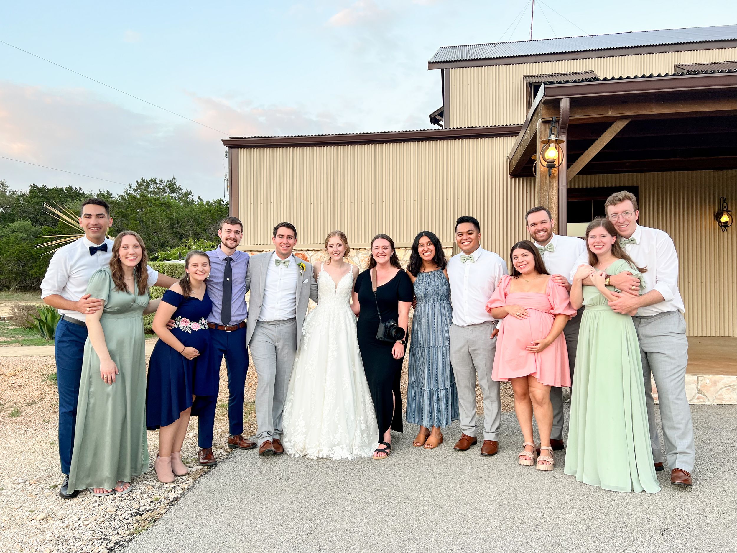 Wedding party in pastel attire poses outside rustic venue at sunset.