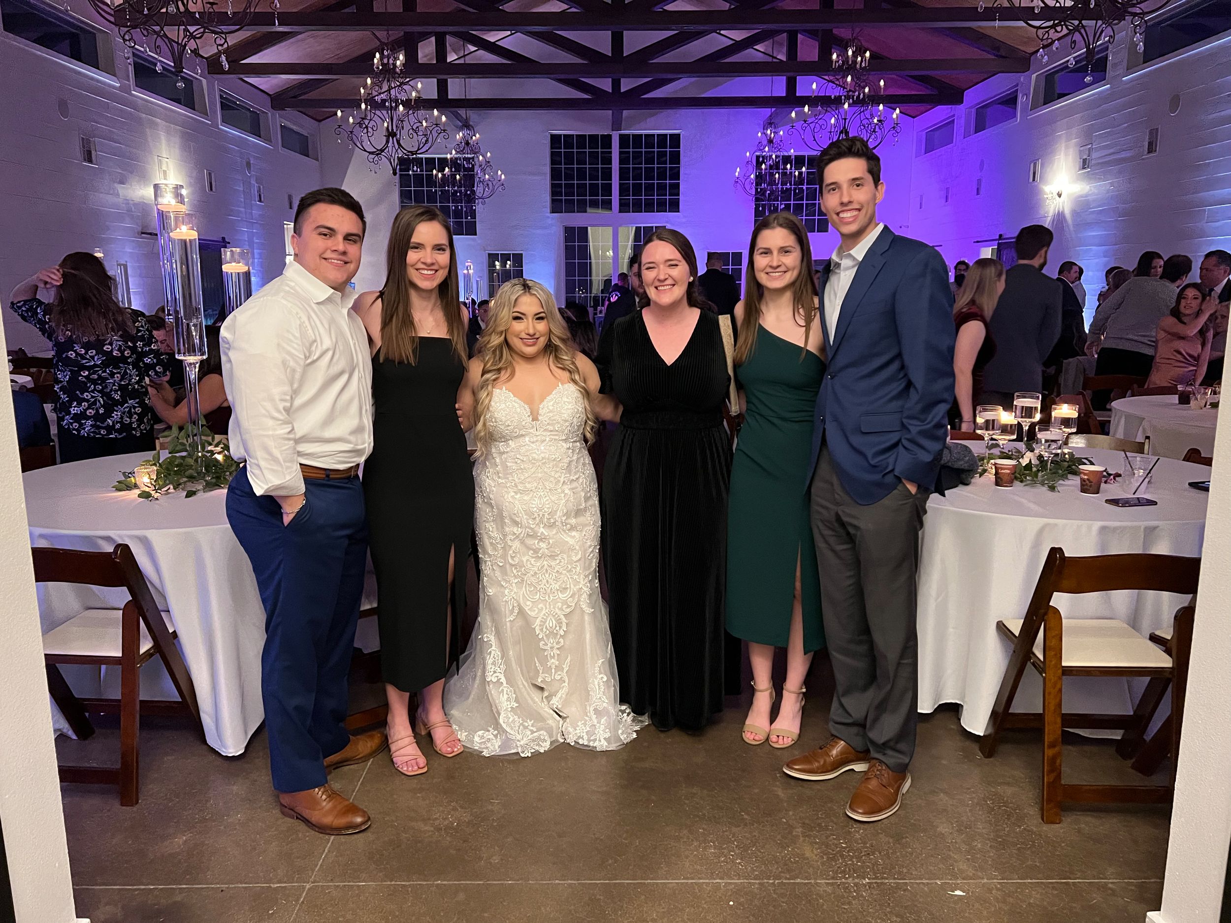 Wedding guests dance and celebrate on an illuminated dance floor at an indoor reception venue with purple lighting.