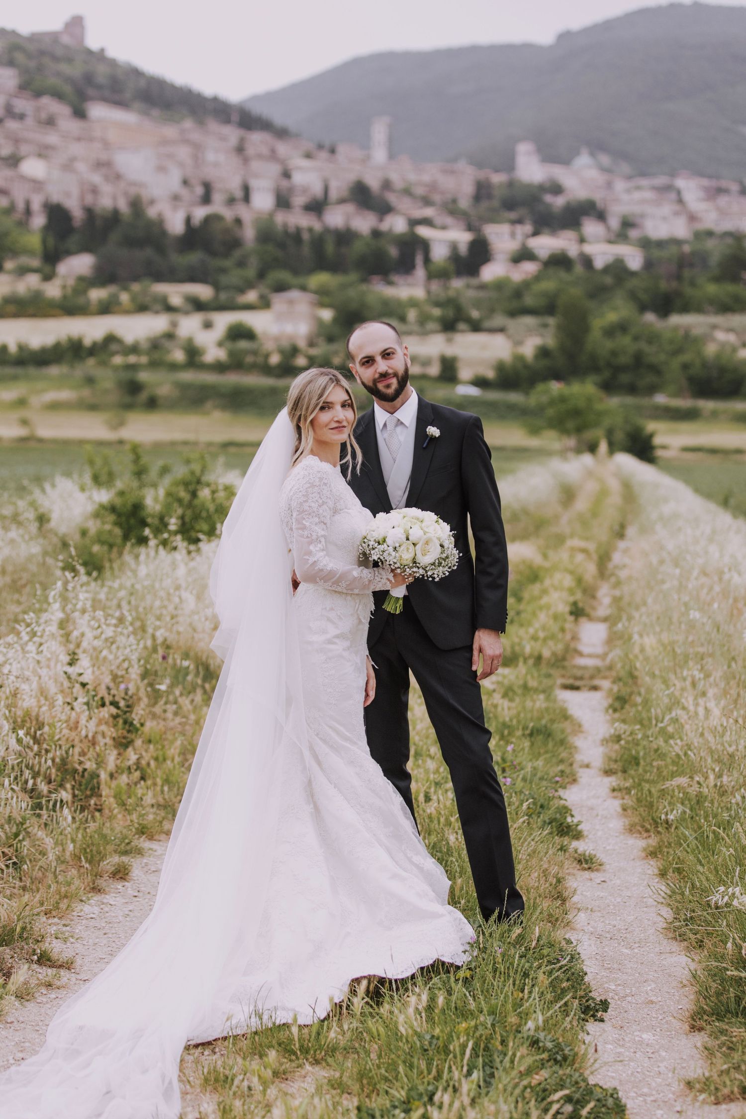 A wedding couple walks along a path through golden meadows with a hilltop town vista.