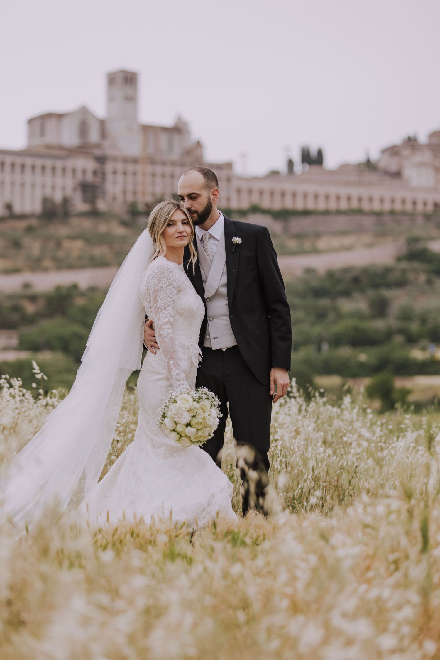 Newlyweds stand in a wildflower field with a historic Italian town and basilica in the background.
