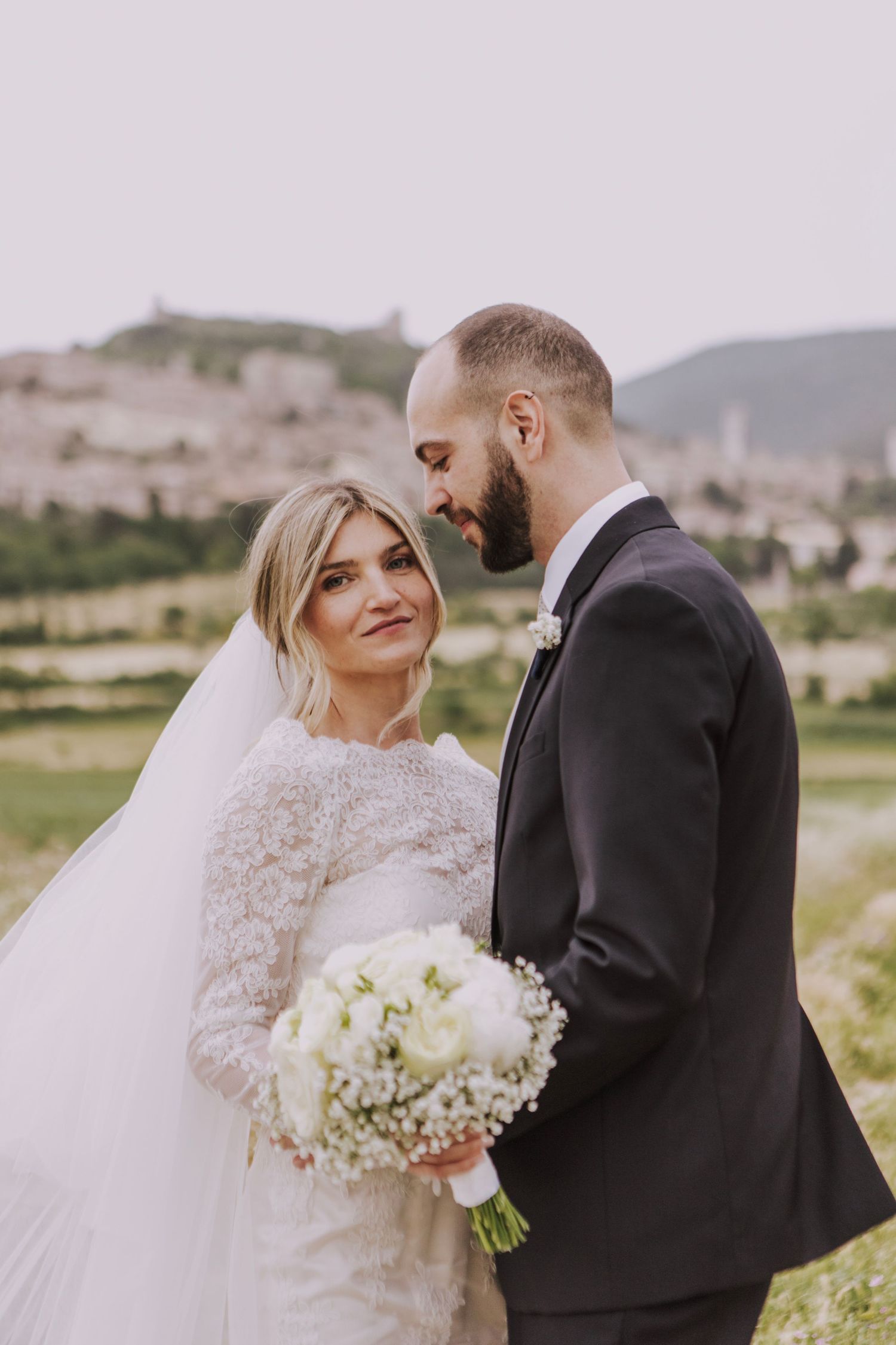 A tender moment between newlyweds holding a white bouquet against a scenic Italian landscape.