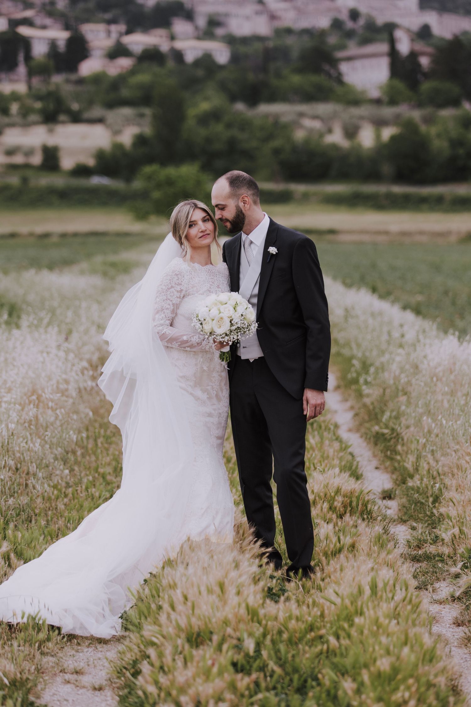 A couple in wedding attire poses together in a field with distant buildings in the background.