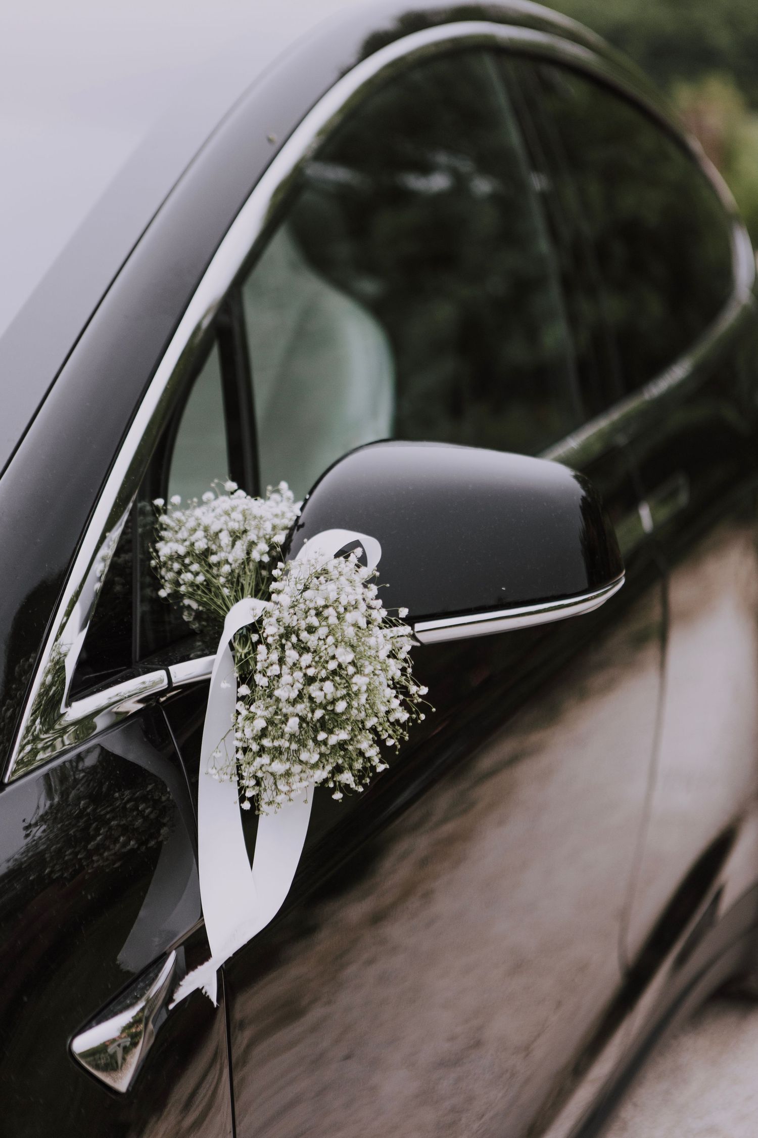 White baby's breath flowers and ribbons decorate the side mirror of a black wedding car.