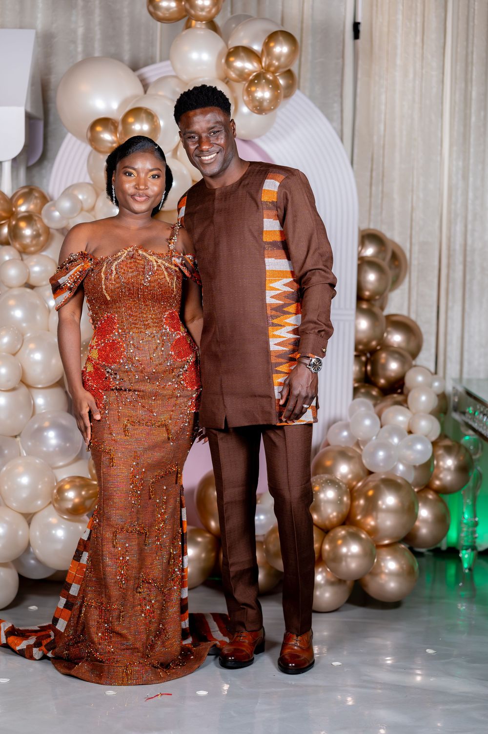 Couple in formal attire posing together against a backdrop of gold and white balloons at an event.
