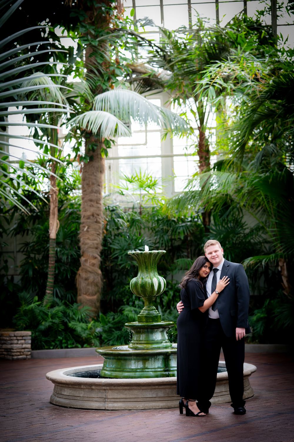 Romantic engagement photo session in a lush indoor garden conservatory with a decorative fountain.