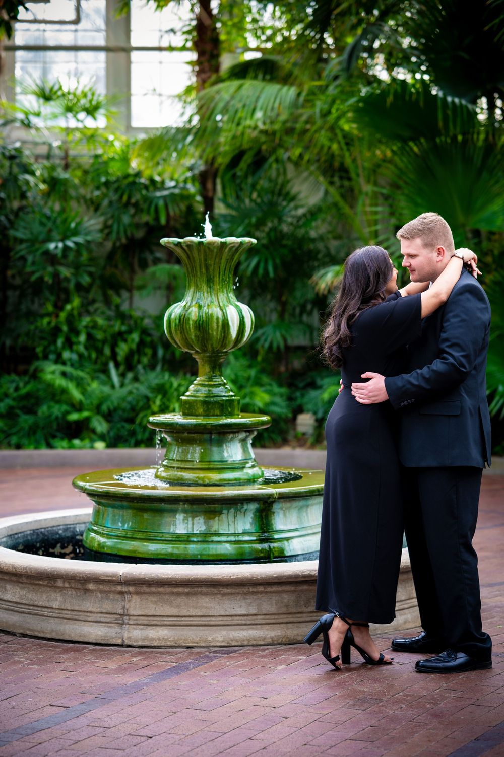 Couple poses near an ornate green fountain in a tropical indoor garden setting.