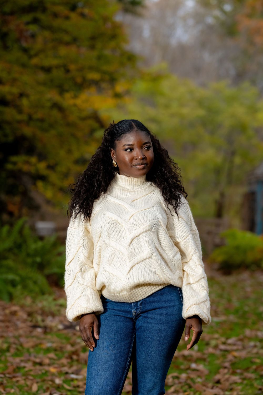 Person in cream cable knit sweater and jeans poses among fallen autumn leaves.