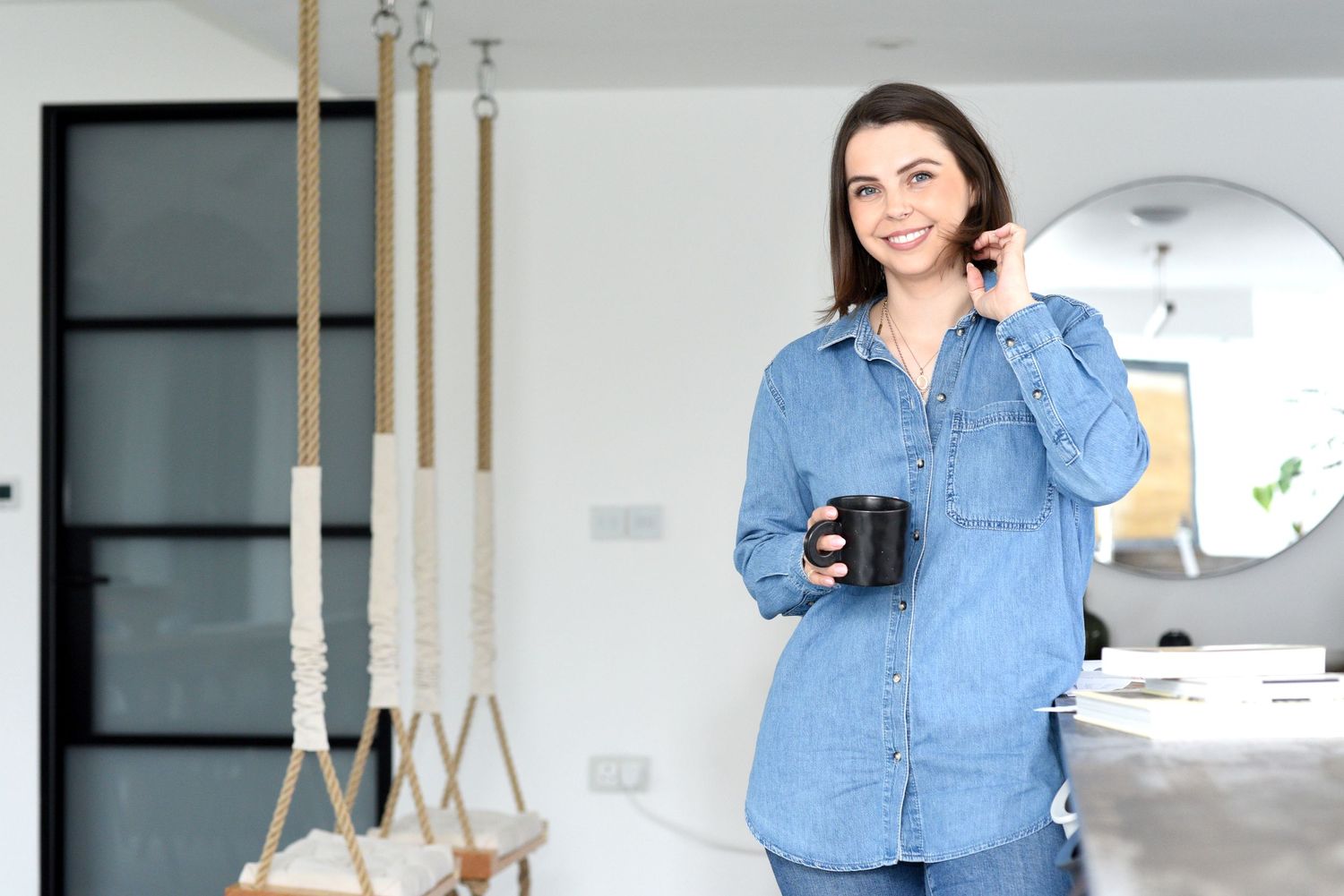 Someone wearing a casual denim shirt holds a black coffee mug while standing in a modern white interior space. Taken on a brand photoshoot.