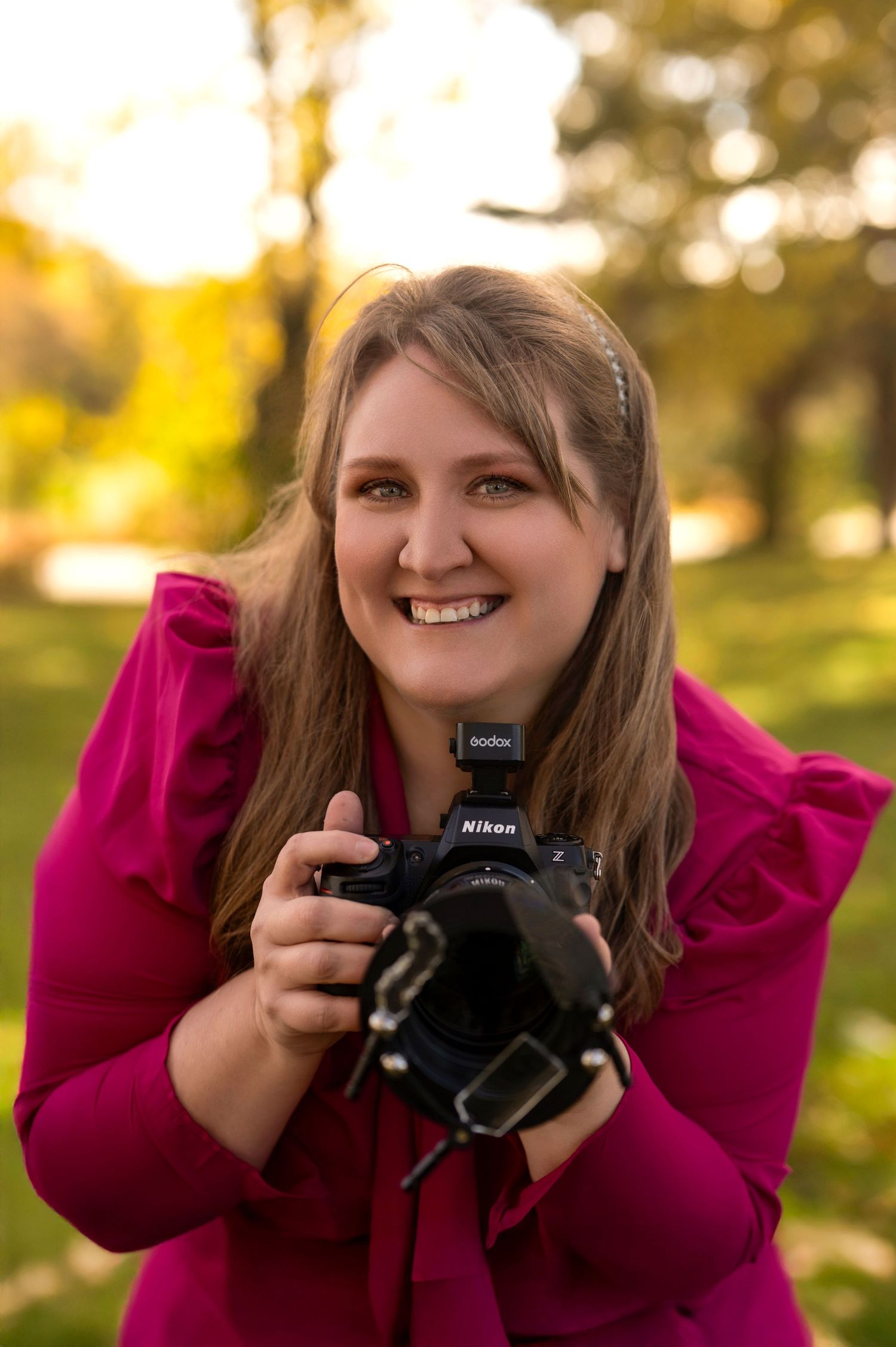 Smiling female photographer holding a Nikon camera with flash in a sunlit autumn park.