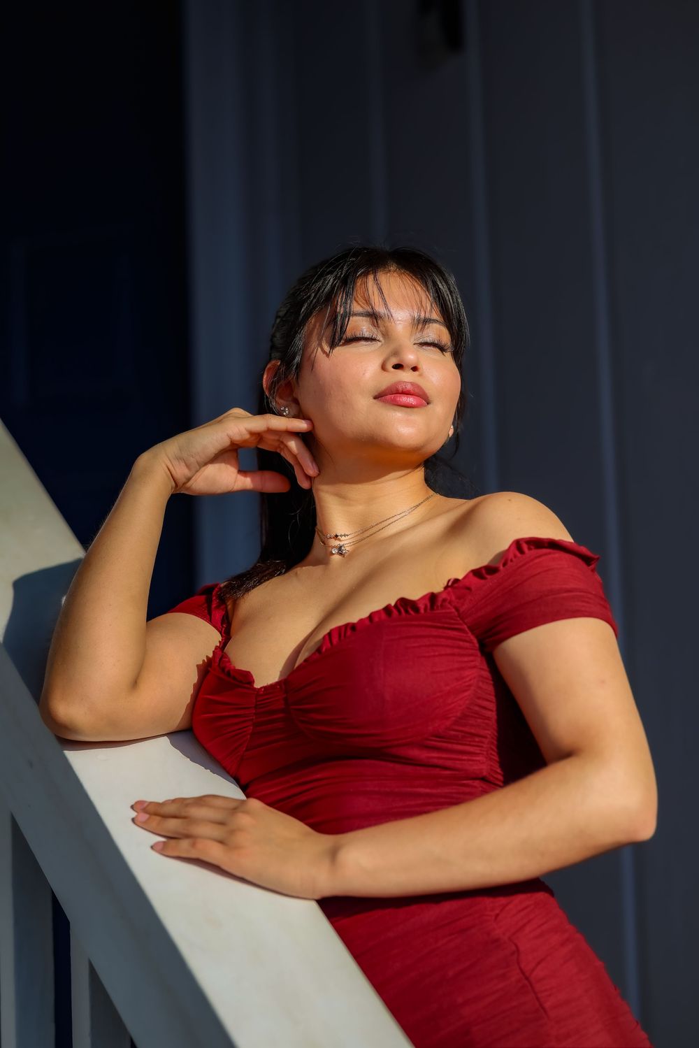 Young woman in red off-shoulder dress leaning on white railing, eyes closed, basking in sunlight.
