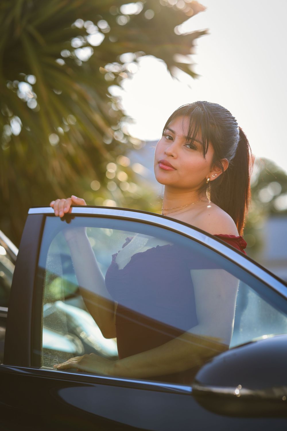 Young woman with ponytail standing by open car door in golden sunlight with palm trees in background.