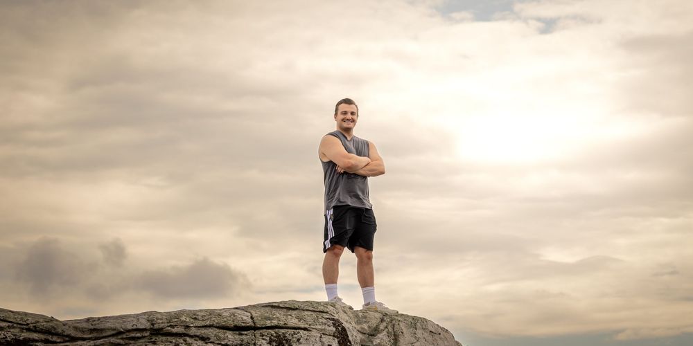 Confident fit man standing on rocky summit with arms crossed, wearing athletic wear against dramatic cloudy sky.