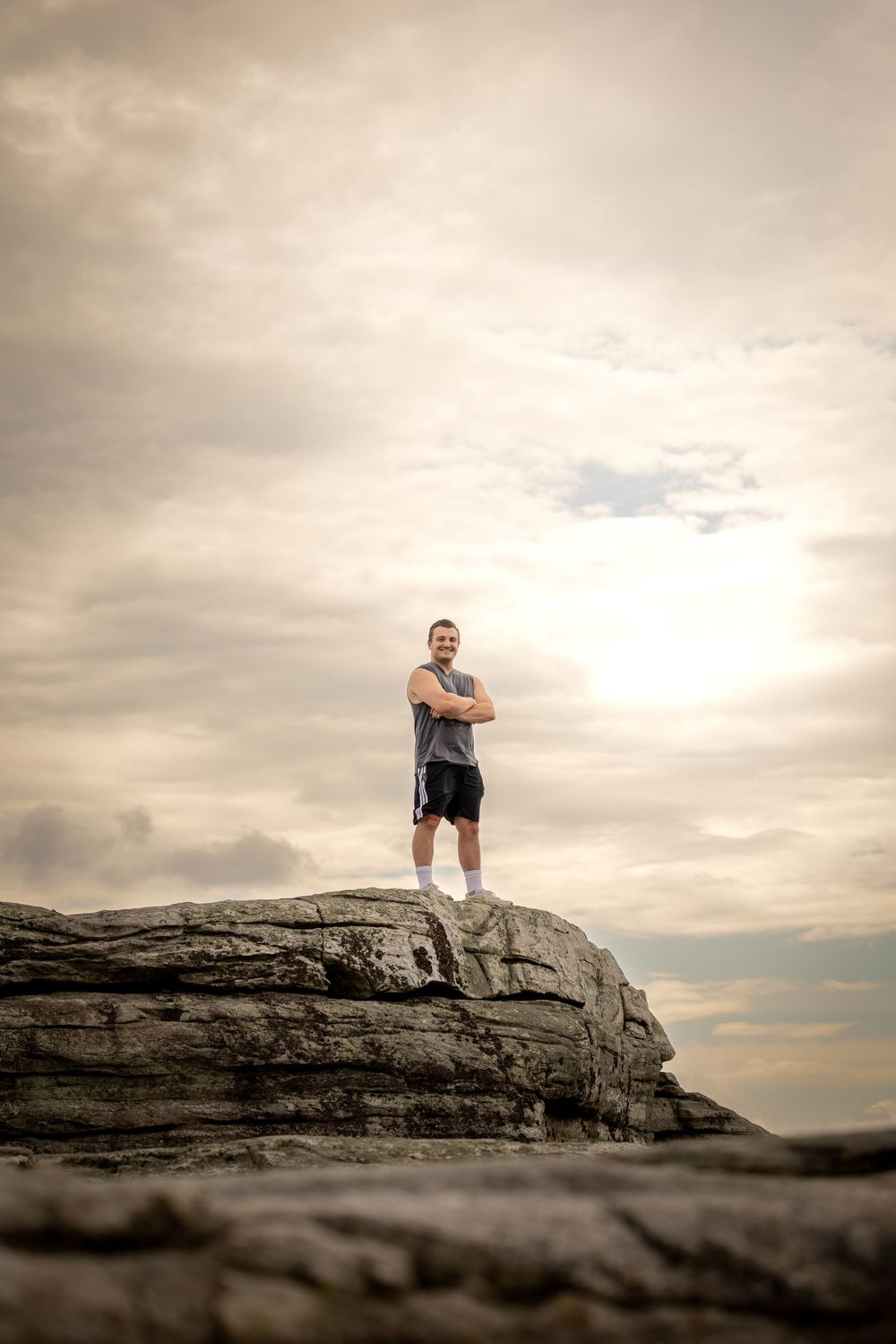 A confident man stands arms crossed on a rocky summit against a dramatic cloudy sky at sunset.