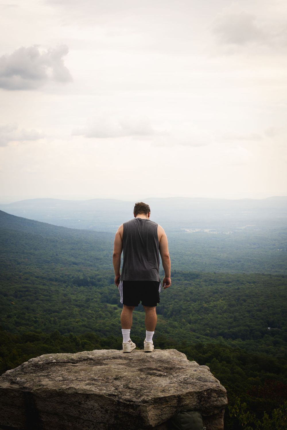Man standing on rocky cliff edge looking down at expansive green mountain landscape below.