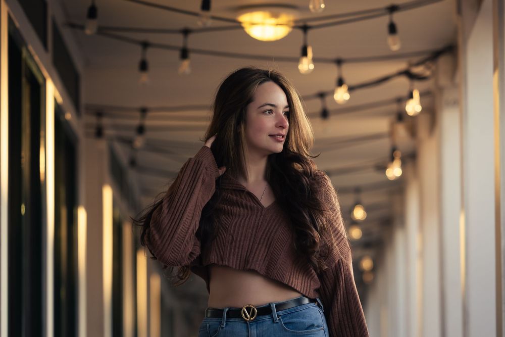 Young woman in brown crop sweater and jeans poses in warmly lit corridor with string lights overhead.