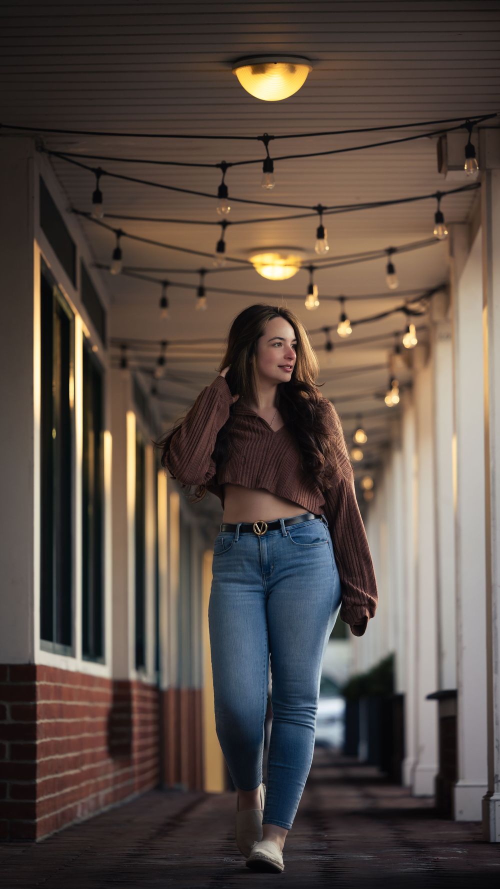 Young woman in jeans and brown sweater walks along warmly lit corridor with string lights overhead.