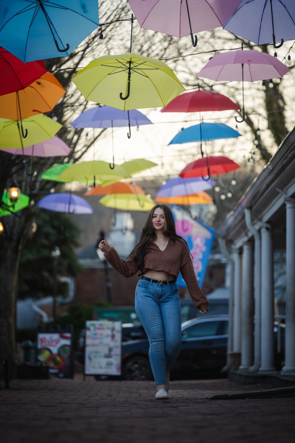 Young woman walks under colorful hanging umbrellas on a charming outdoor street, wearing jeans and brown top.