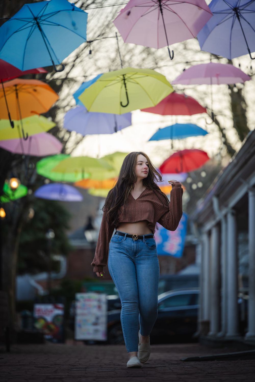 Young woman in brown crop top and jeans walks under colorful hanging umbrellas on a charming street.