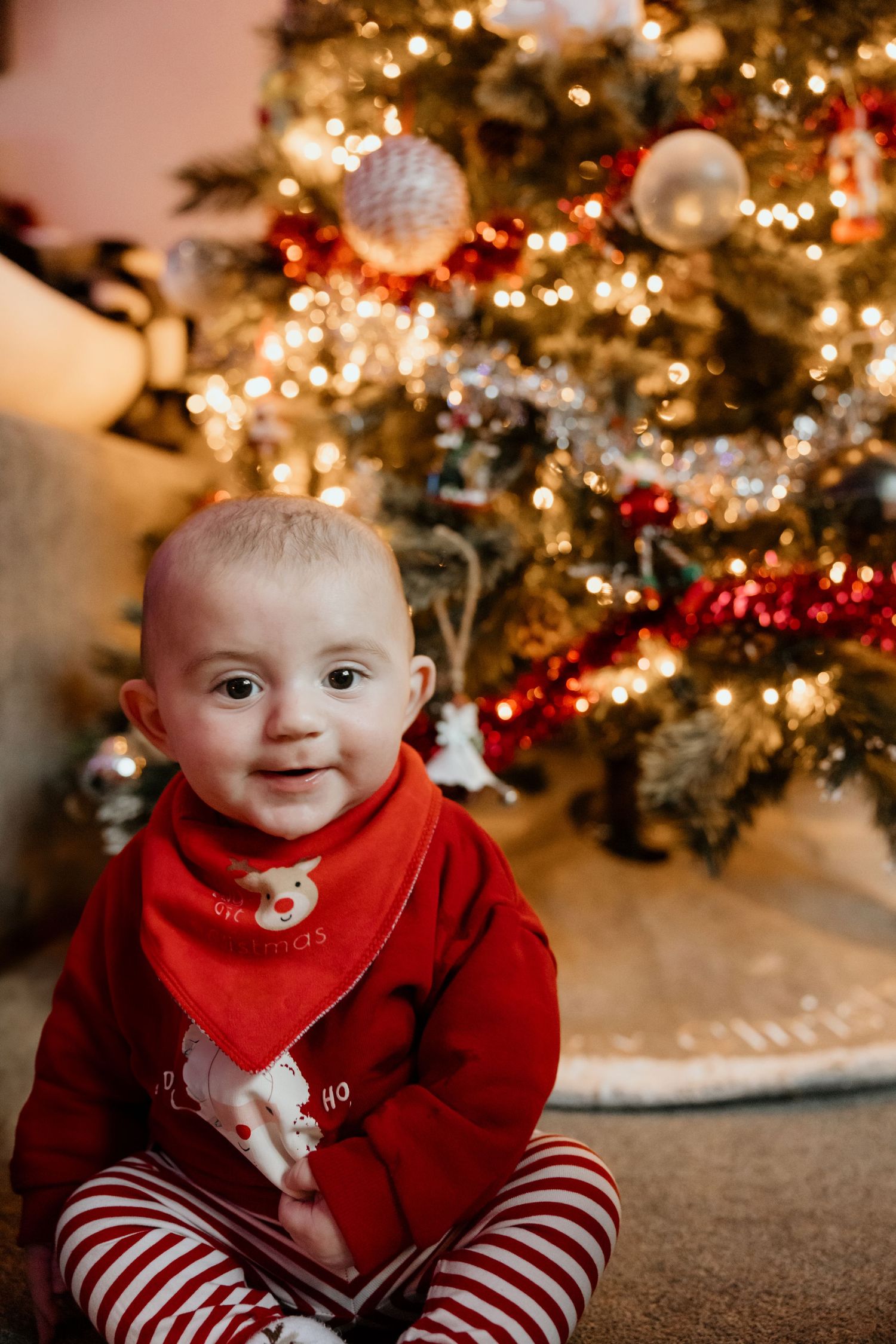 Festive holiday scene with decorated Christmas tree and smiling baby in red and white striped outfit.