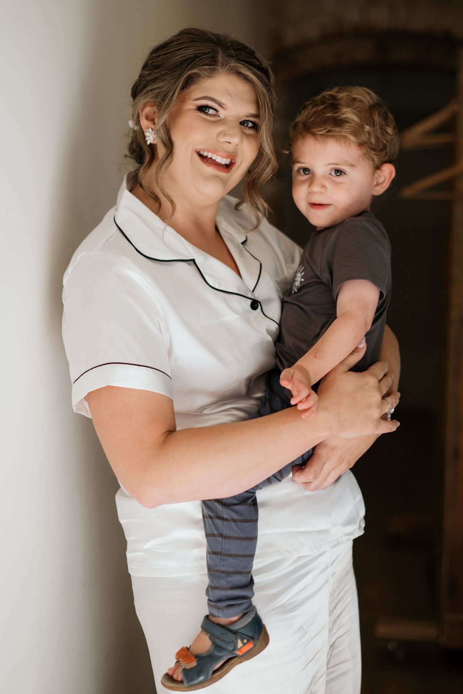 A smiling woman in white pajamas holds a toddler boy in a gray shirt and blue pants, posing indoors.