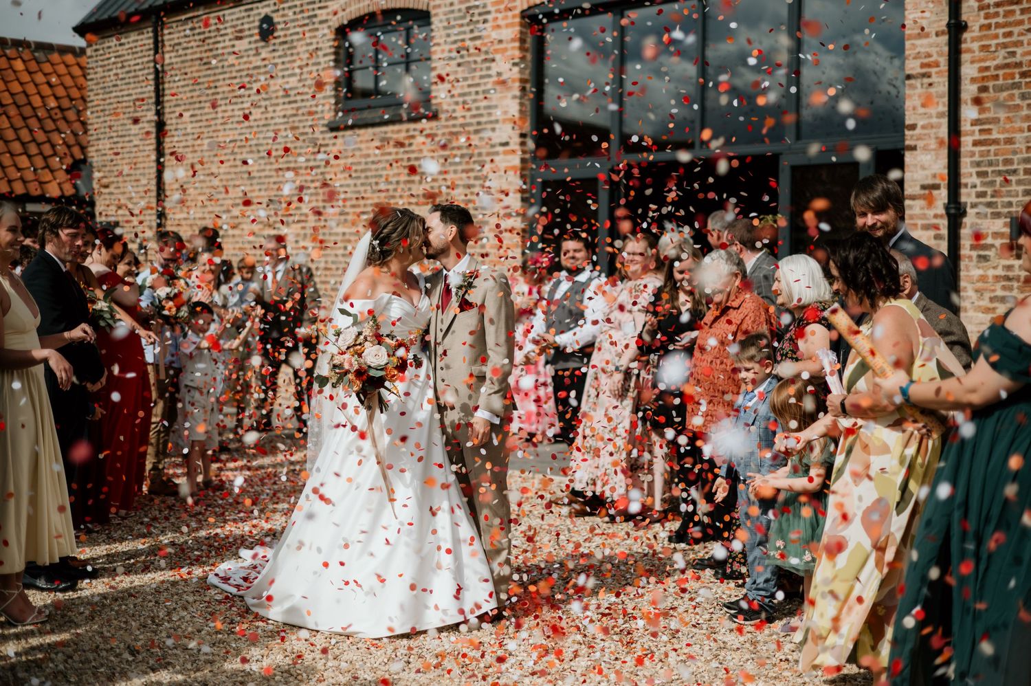 Bride and groom kissing under colorful confetti shower outside rustic brick venue surrounded by wedding guests.