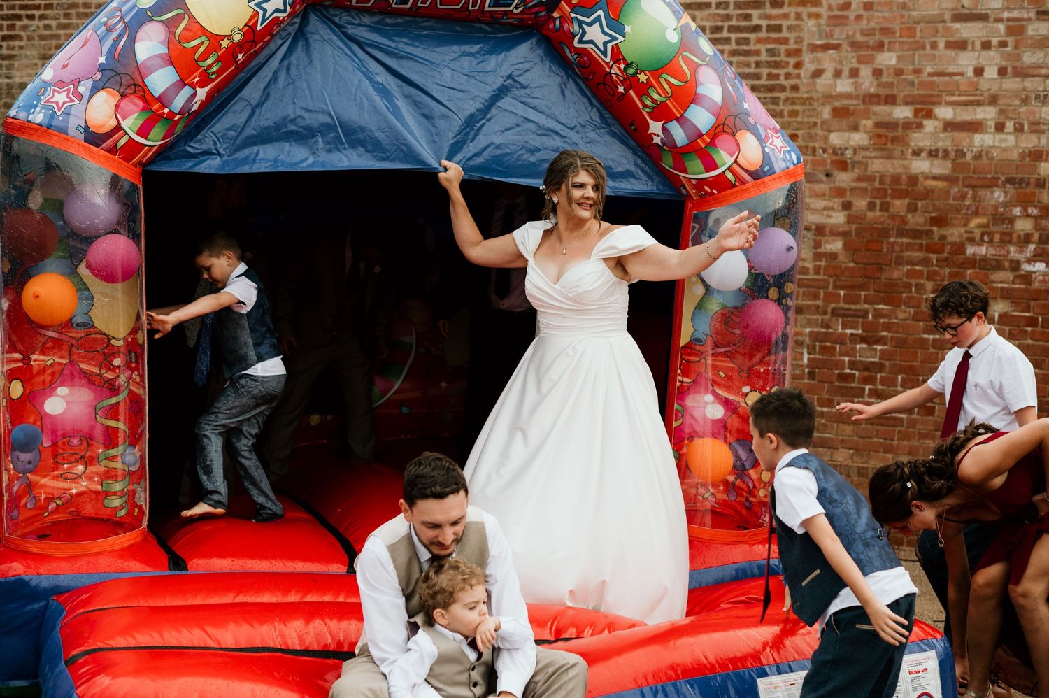 Bride in white gown laughing on colorful bouncy castle with wedding guests at outdoor reception.