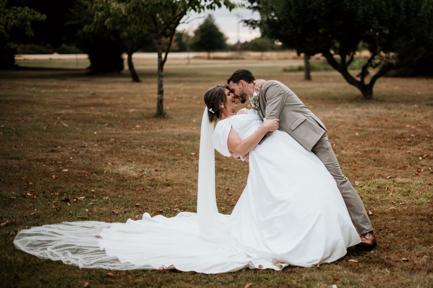 Groom dips bride in romantic outdoor wedding kiss, white gown with long veil trailing on green grass among trees.