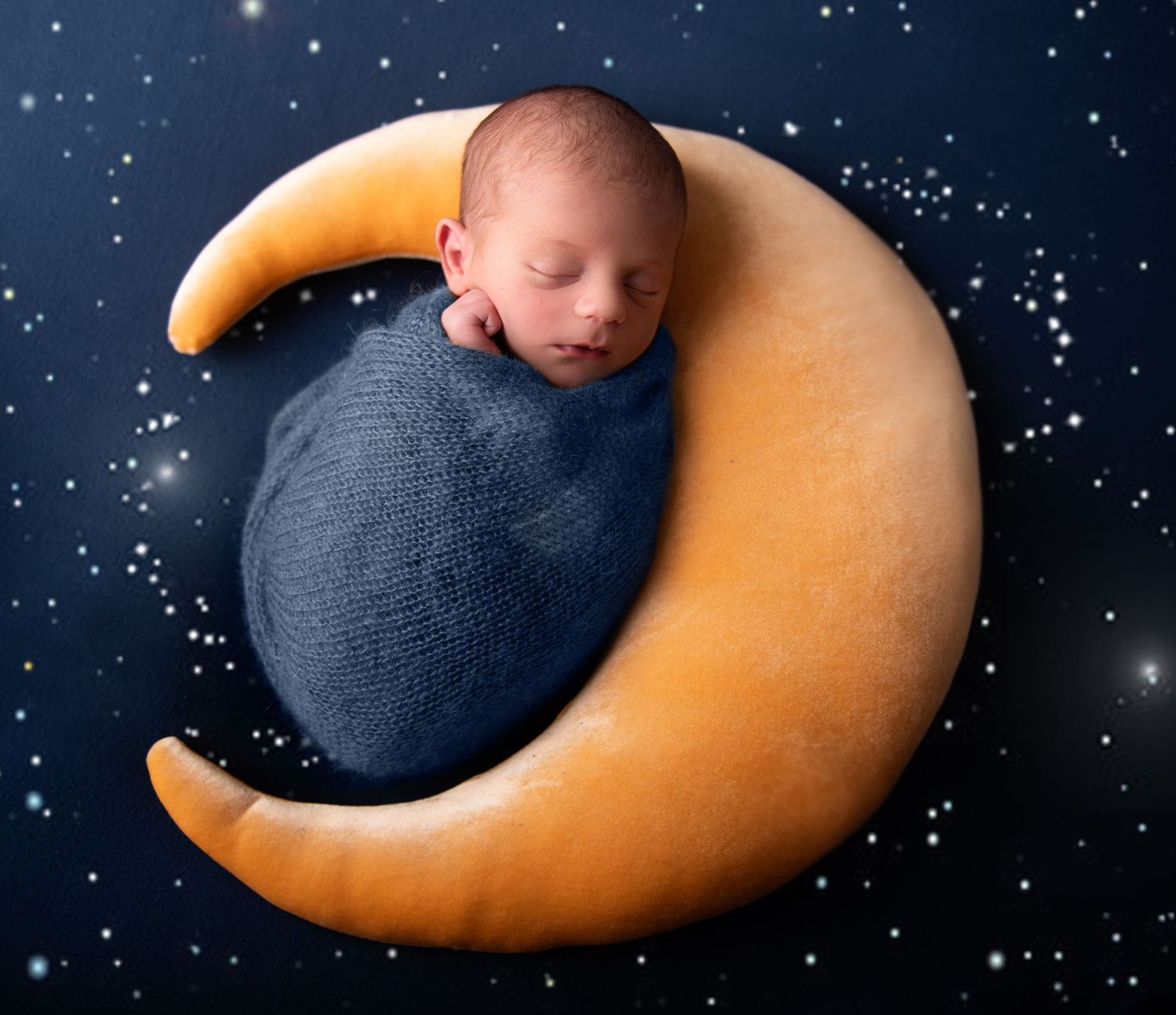 Newborn baby wrapped in navy blue swaddle sleeping peacefully on decorative crescent moon prop against starry background.