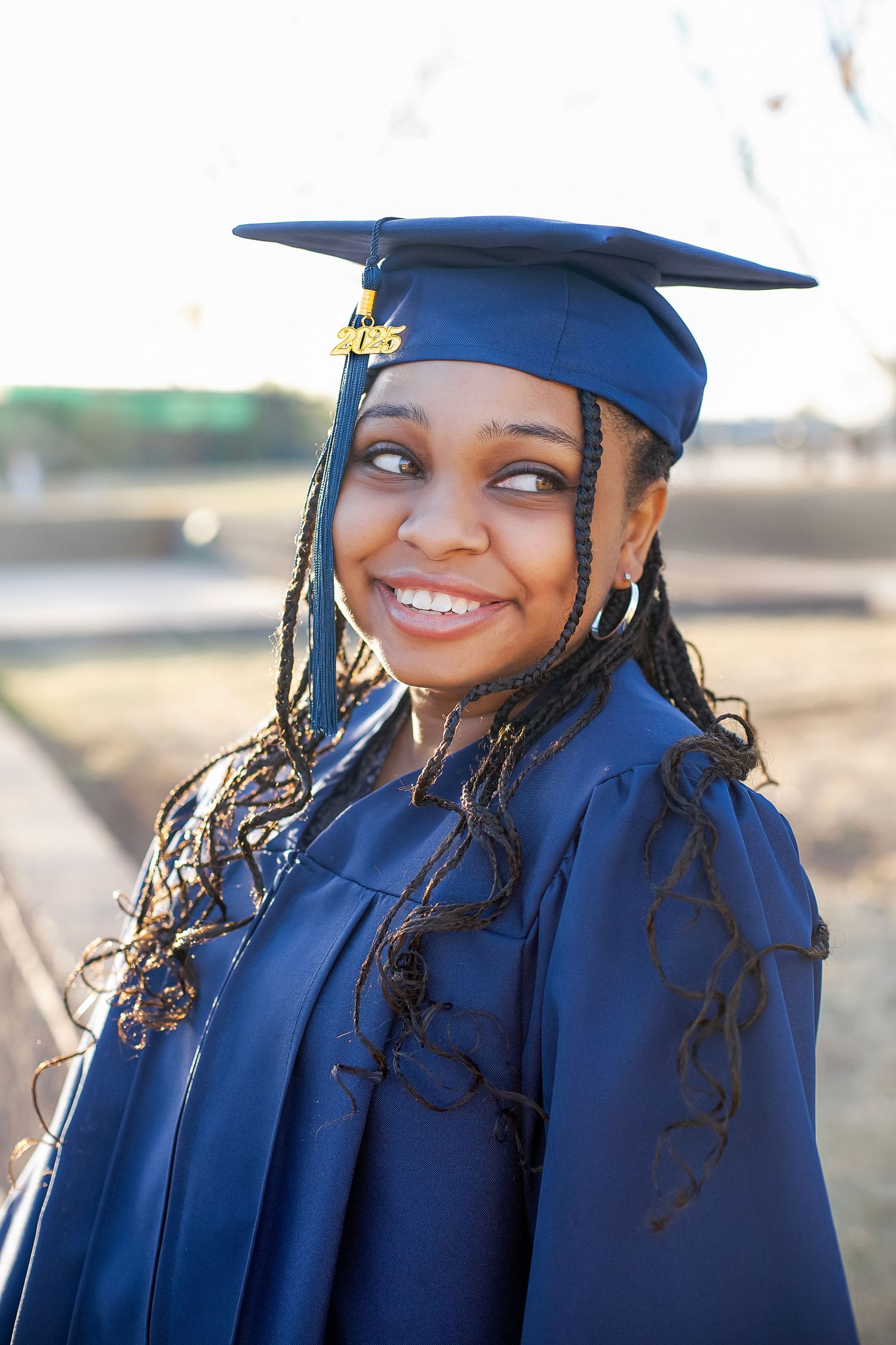 Smiling young Black woman in blue graduation cap and gown with long braids, celebrating her academic achievement outdoors.