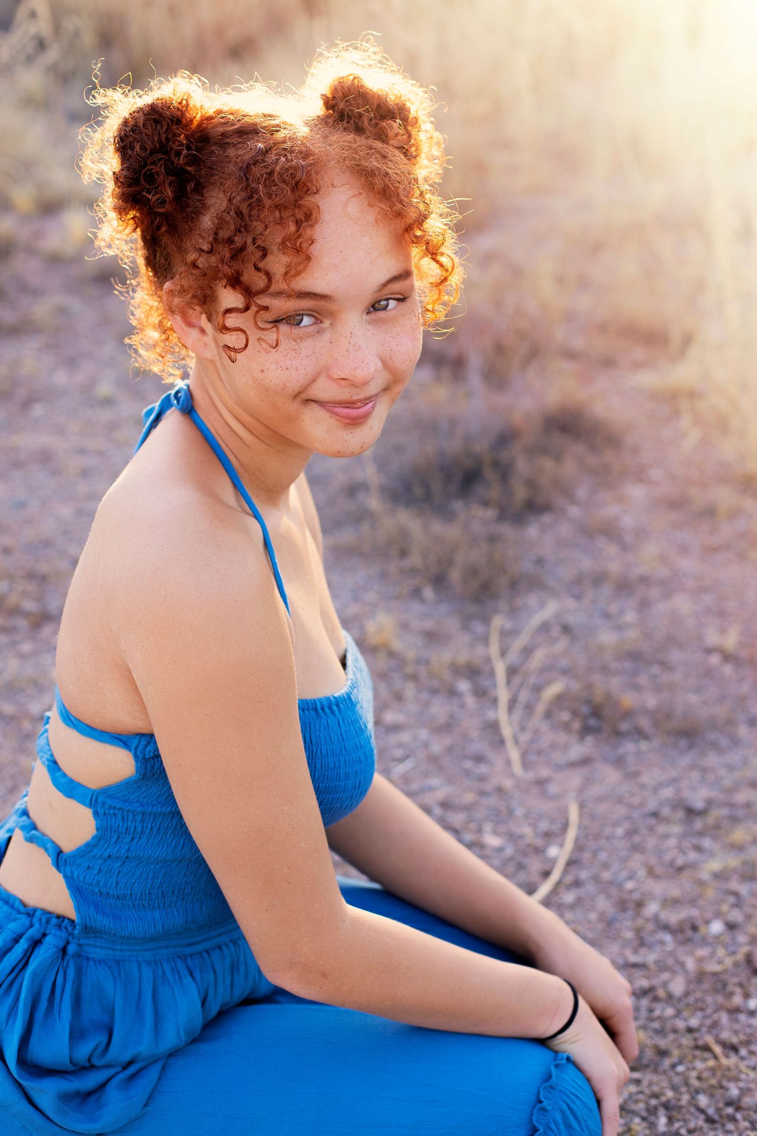 Young redhead girl with curly hair in blue dress sits outdoors in sunlit desert landscape, smiling over shoulder.