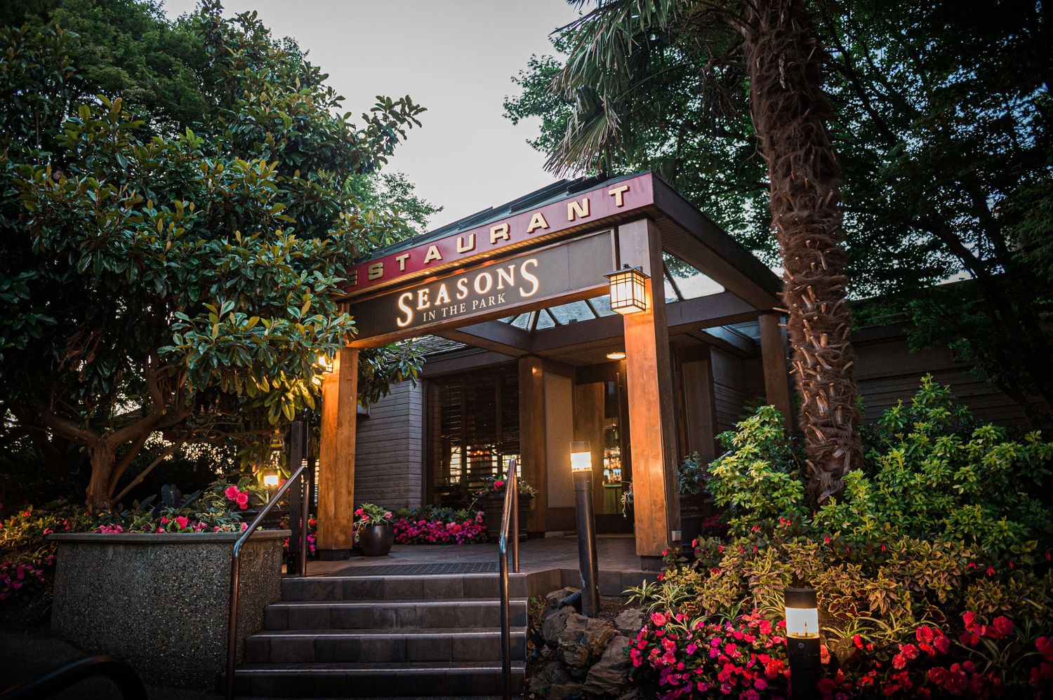 Illuminated restaurant entrance with 'Seasons' sign surrounded by colorful flower planters at dusk.