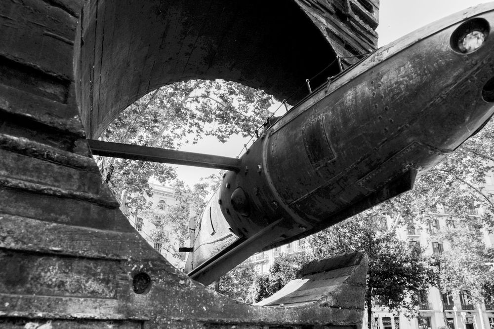 Black and white photo of vintage wooden boat hull with torpedo-shaped object mounted underneath, surrounded by trees.