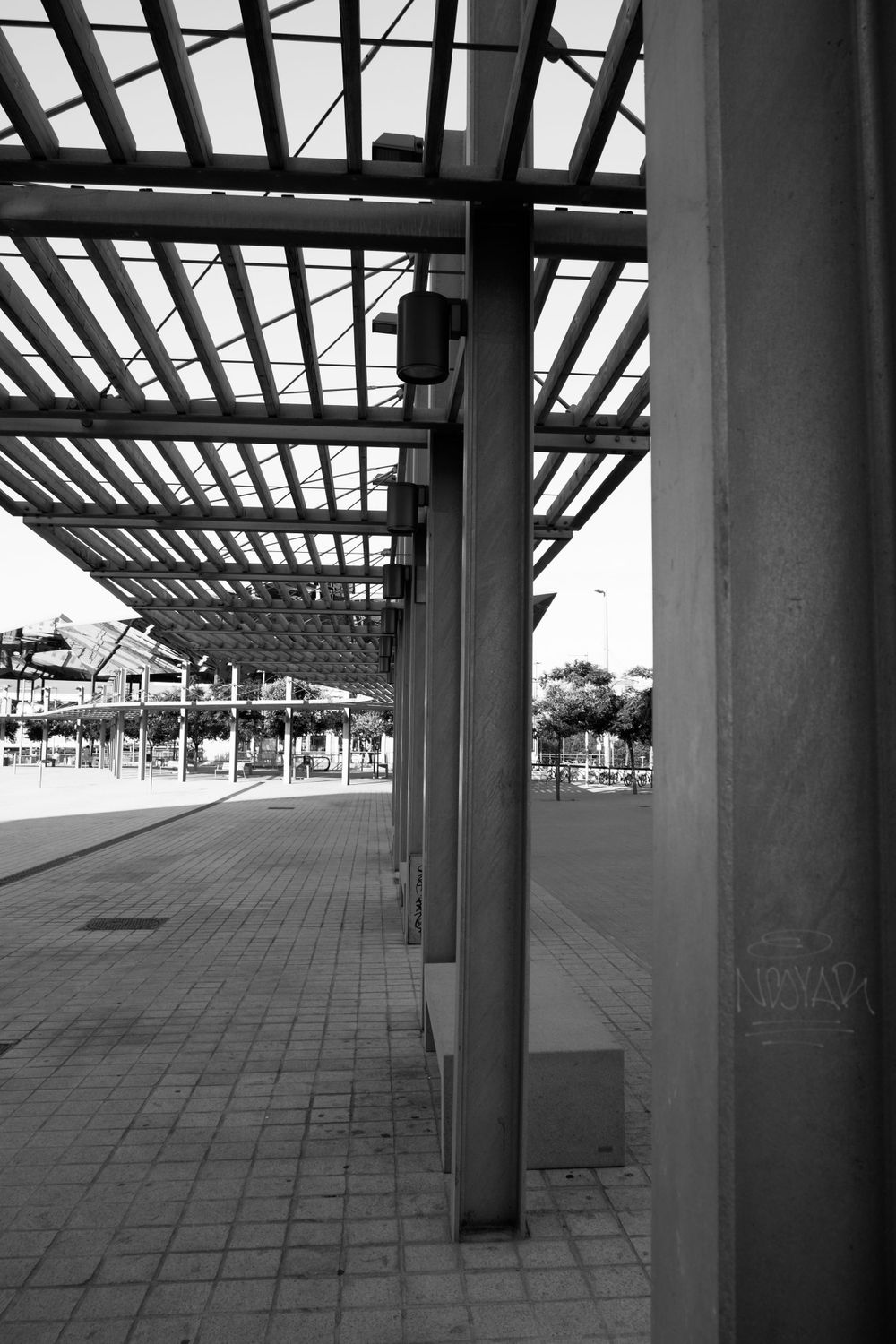 Black and white photo of empty covered walkway with columns and geometric pergola roof.