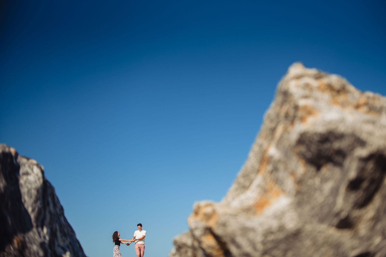 Two figures stand atop a rocky mountain peak against a bright blue sky.