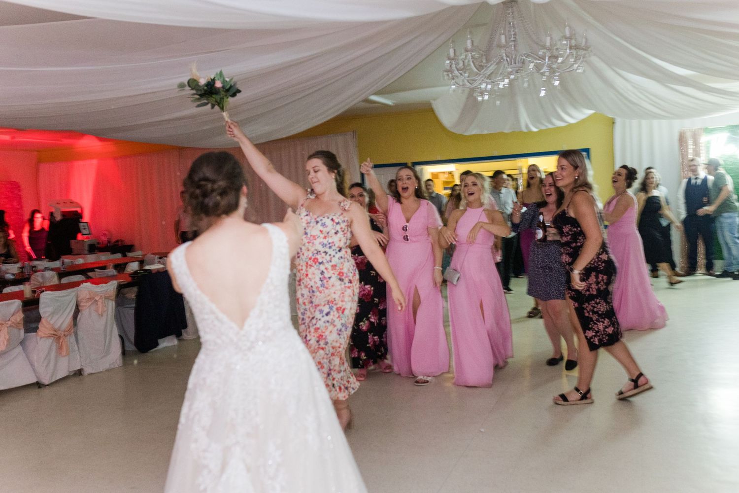 Guests celebrating at a wedding reception with pink lighting and white draped ceiling decor.