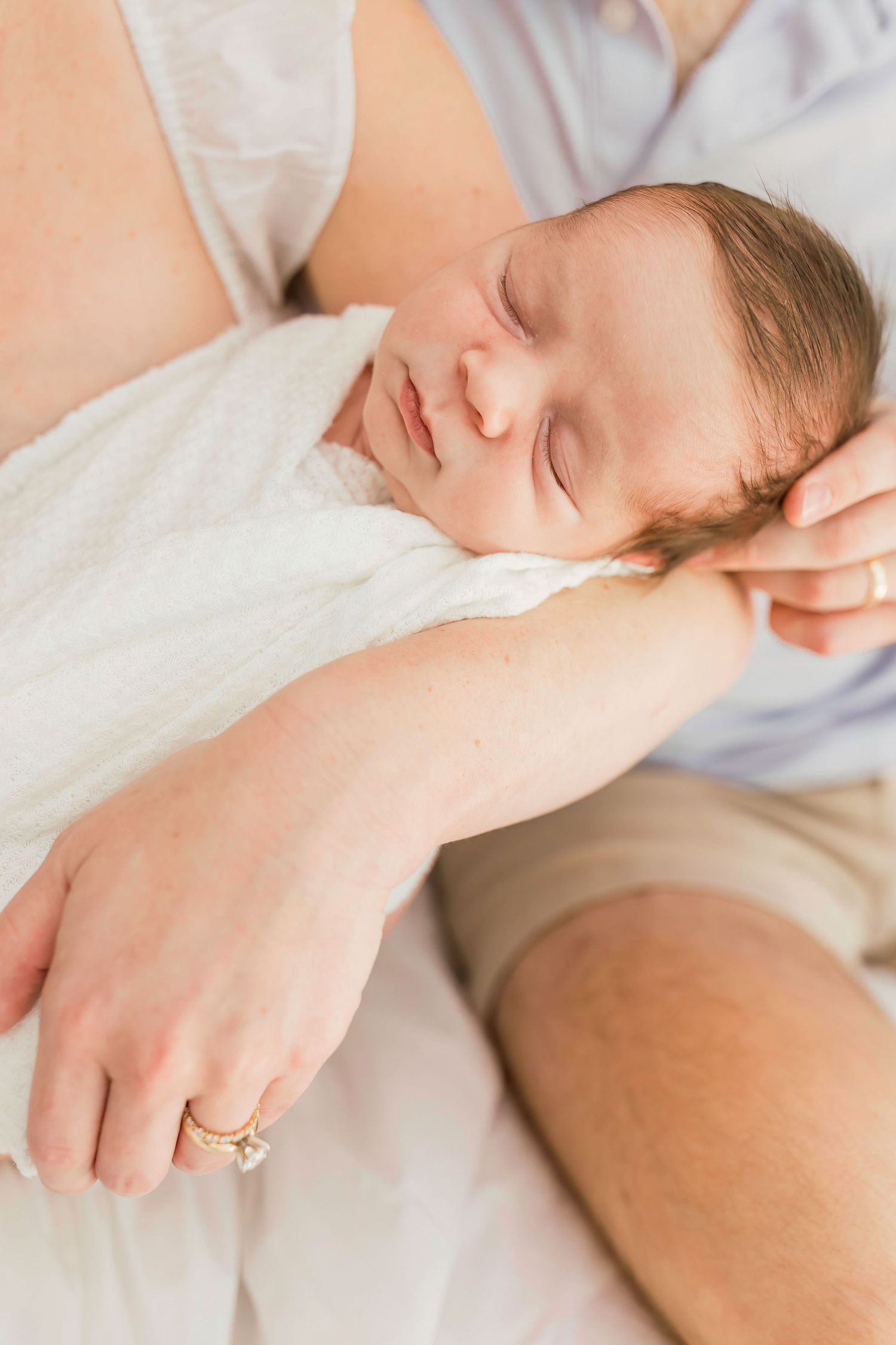 A sleeping newborn baby wrapped in white with parent's hand showing wedding ring.