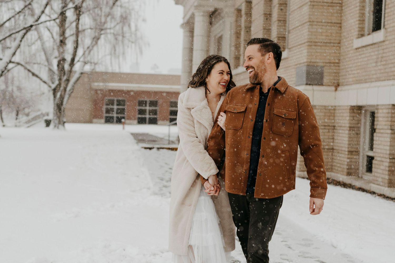 Romantic winter couple walking together in snow outside brick building.