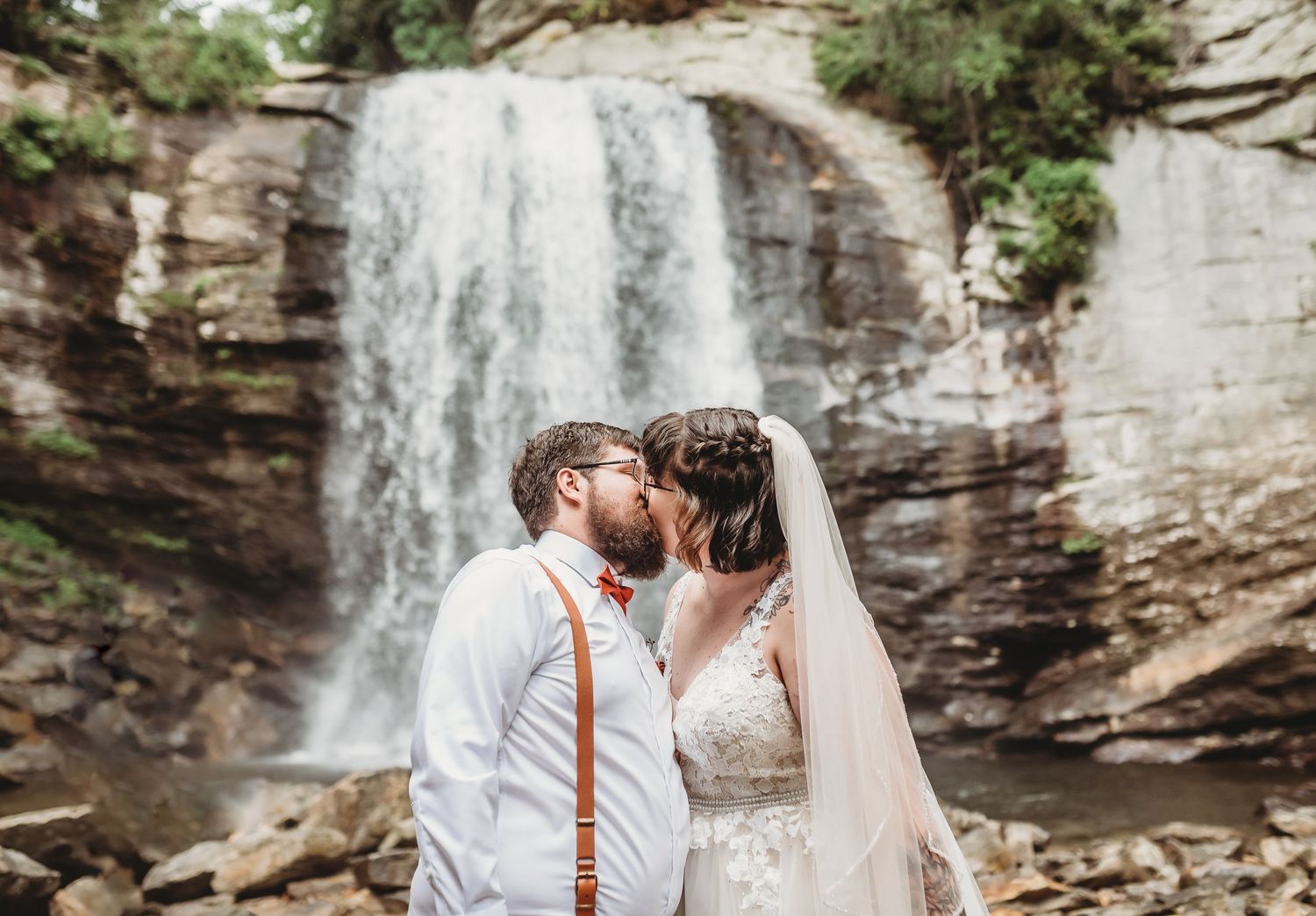 Man and woman in wedding dress kiss in front of waterfall during an elopement in Asheville, NC.