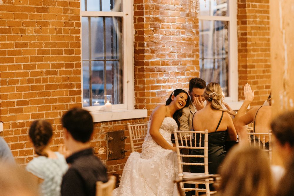 Bride in lace gown shares emotional moment with seated guest at wedding reception.